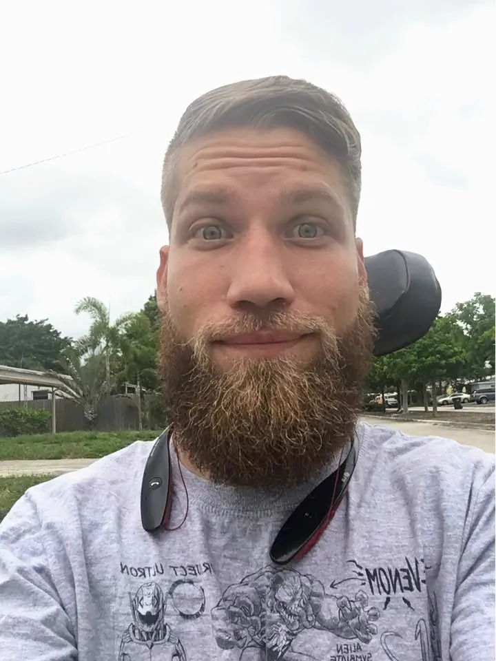 Selfie of Jimmy outdoors, wide-eyed with a half smile and a big reddish beard, wearing a gray graphic T-shirt with headphones around his neck.