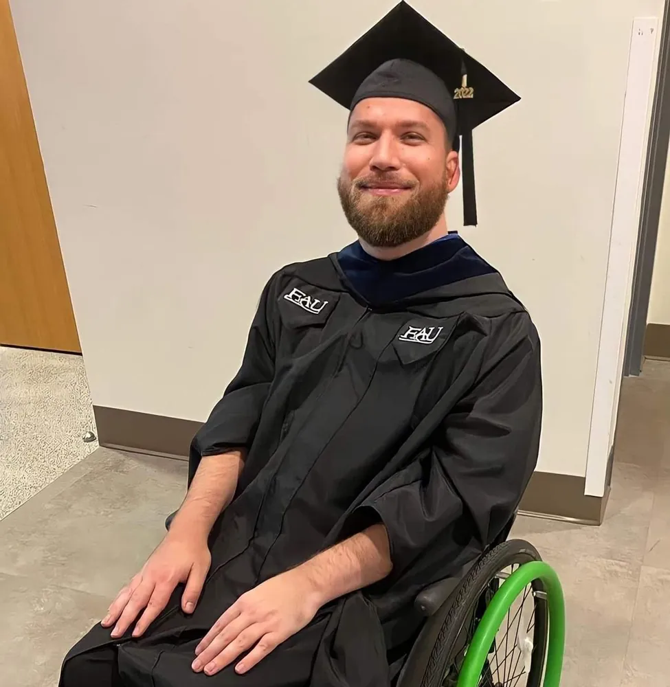 James smiling in a black Florida Atlantic University cap and gown with a 2022 tassel, seated in his wheelchair.